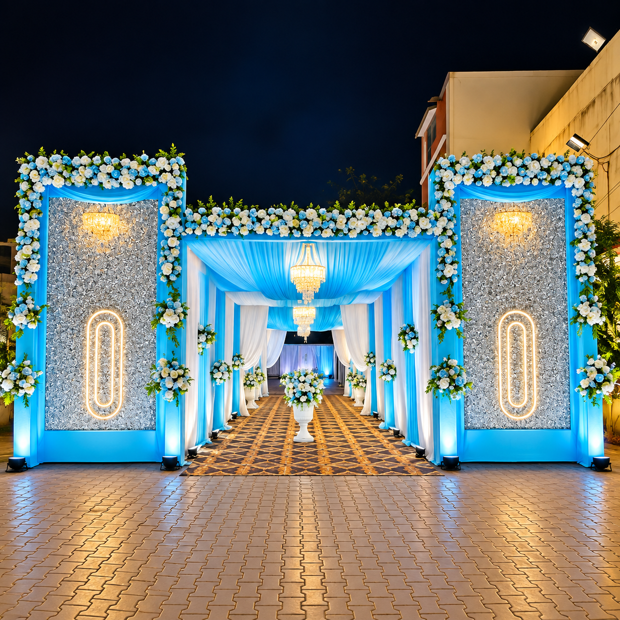 Outdoor wedding entrance at night with blue and white drapes, flower garlands, chandeliers and silver sequin panels